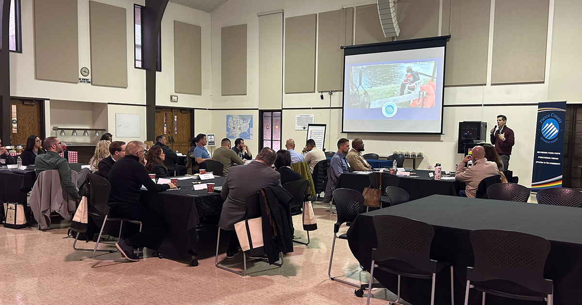 A large group of people sit together for a presentation. They watch a large screen on which a video plays.