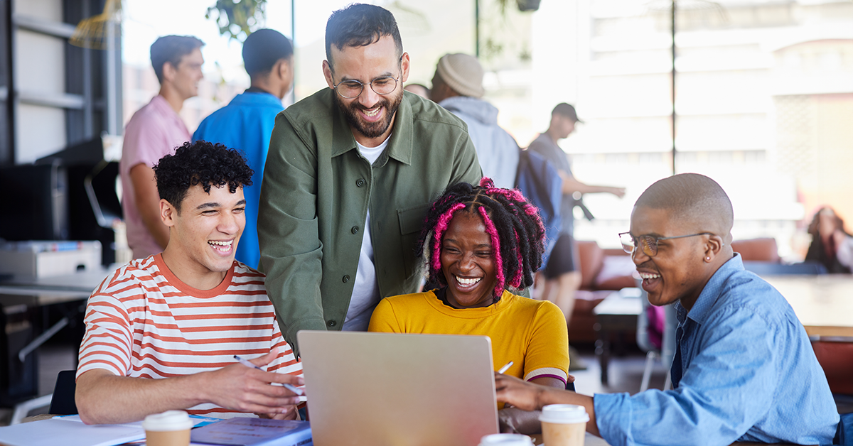 A group of four young people stand around a laptop, talking happily, in a communal space.
