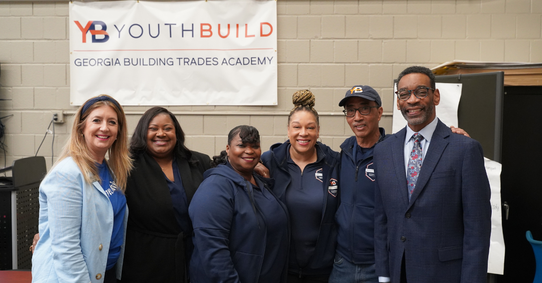 Group of six adults standing together and smiling in front of a “YouthBuild Georgia Building Trades Academy” banner, including program participants and a suited representative, inside a training space.