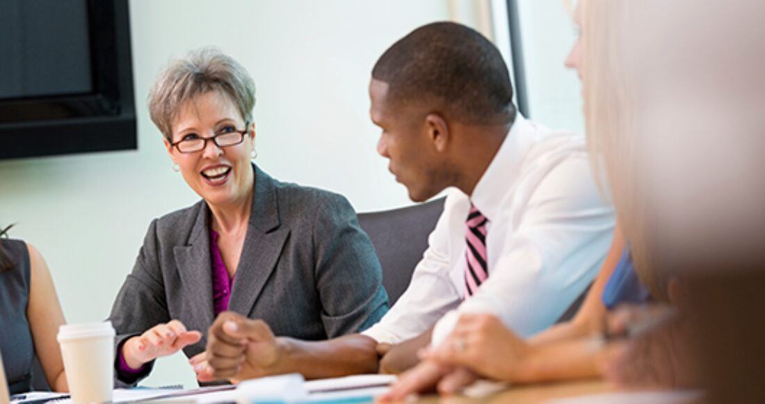 In an office, a woman sits at table smiling to a male colleague sitting next to her.