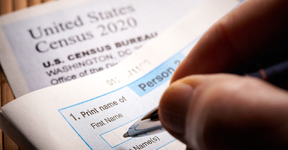 The image is a close-up shot of a hand, holding a pen. The pen hovers above a blank questionnaire. Underneath the questionnaire is a form that says, "United States Census 2020."