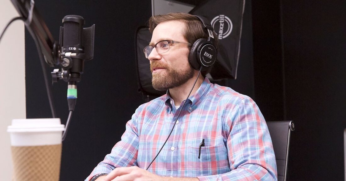 A white man sits in a recording studio, wearing headphones. The man is smiling and wearing glasses. A microphone is in front of him.