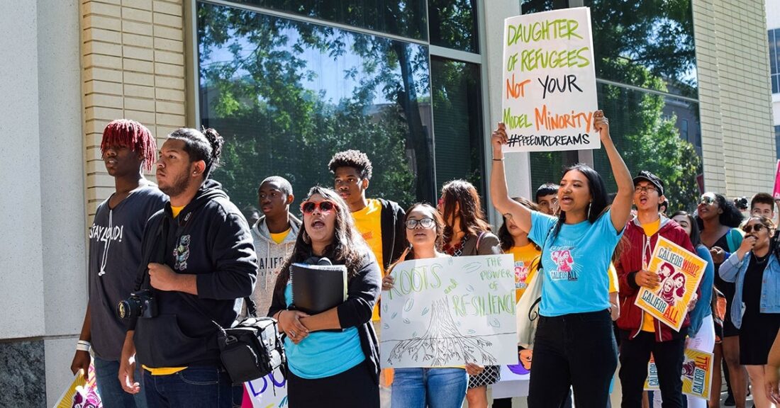 A group of Black and Brown youth gather peacefully outside of a building. Several young men and women hold signs advocating for better treatment of minorities.