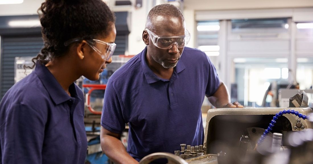 A Black man and a young Brown woman — both wearing goggles and navy blue polo shirts — huddle together over a machine. The man is providing mentorship.