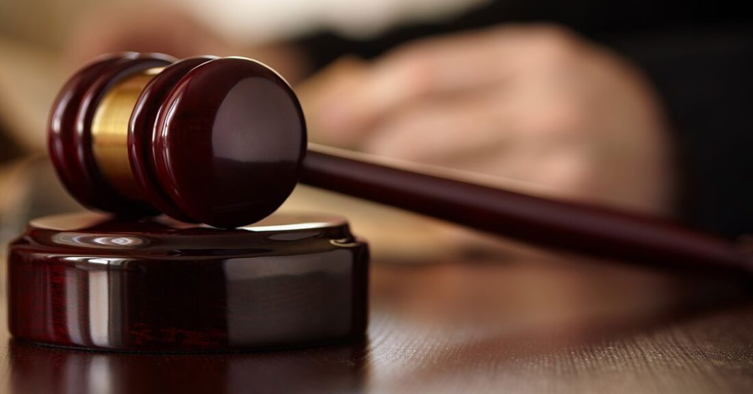 The image is a close-up of a judge's gavel lying on its side on top of its block. A blurry image of the judge's hand is in the background.