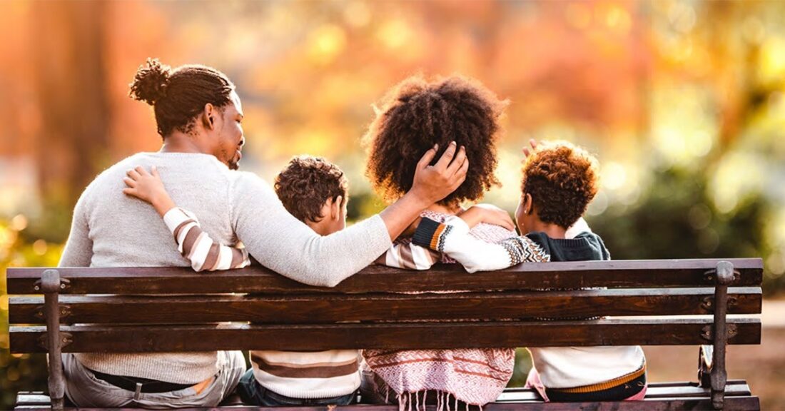 The image is a photo of a Black family of four — taken from behind — sitting  on an outdoor bench, with their arms around each other. The father, on the far left, has dreadlocks pulled into a bun, while the mother has textured, natural hair. They are joined by their two small sons.