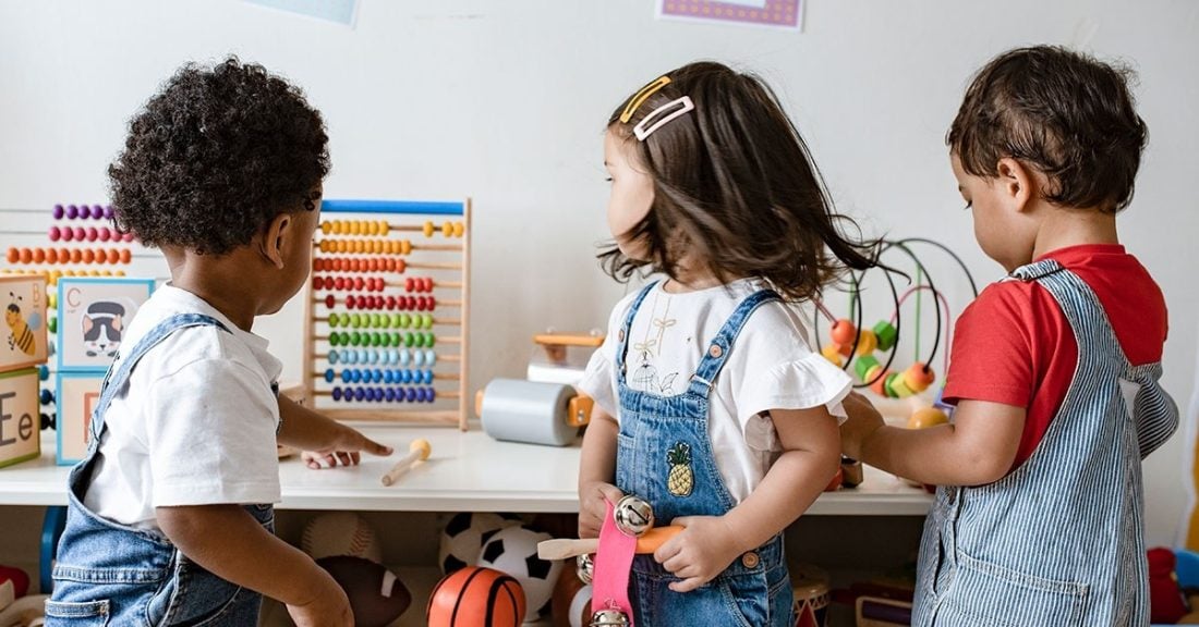 The image depicts three toddlers from behind—a Black boy, a white girl, and a Brown boy—who are in a daycare setting. They are facing a low table on top of which are several colorful learning tools and games.