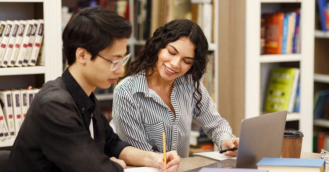 A young Latino woman sits next to an Asian, high-school-aged male student in a library. The young woman, who is smiling, appears to be tutoring the student. A laptop and several notebooks are on the table in front of them.