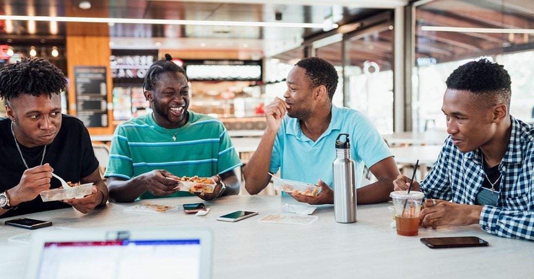 Four college-age, young Black men sit together at a table in a cafeteria. They are smiling, laughing and eating lunch.