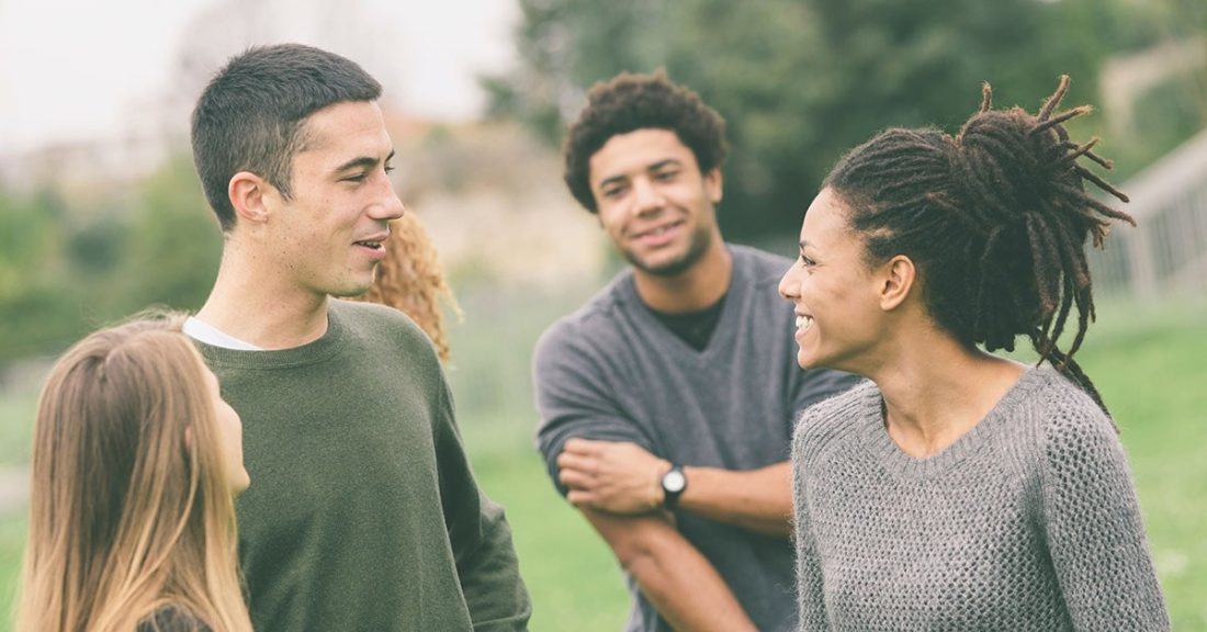 A group of five college-aged young adults gather outdoors. At the forefront of the image is a Black female, with her hair styled in dreadlocks, and a white male. The two are smiling at each other.