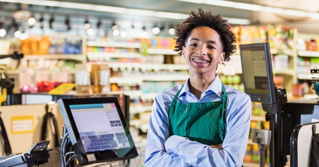 A young, high-school aged Black male, wearing a blue button-down shirt and a green apron, stands behind a cash register while smiling at the camera. He appears to work in a grocery store.