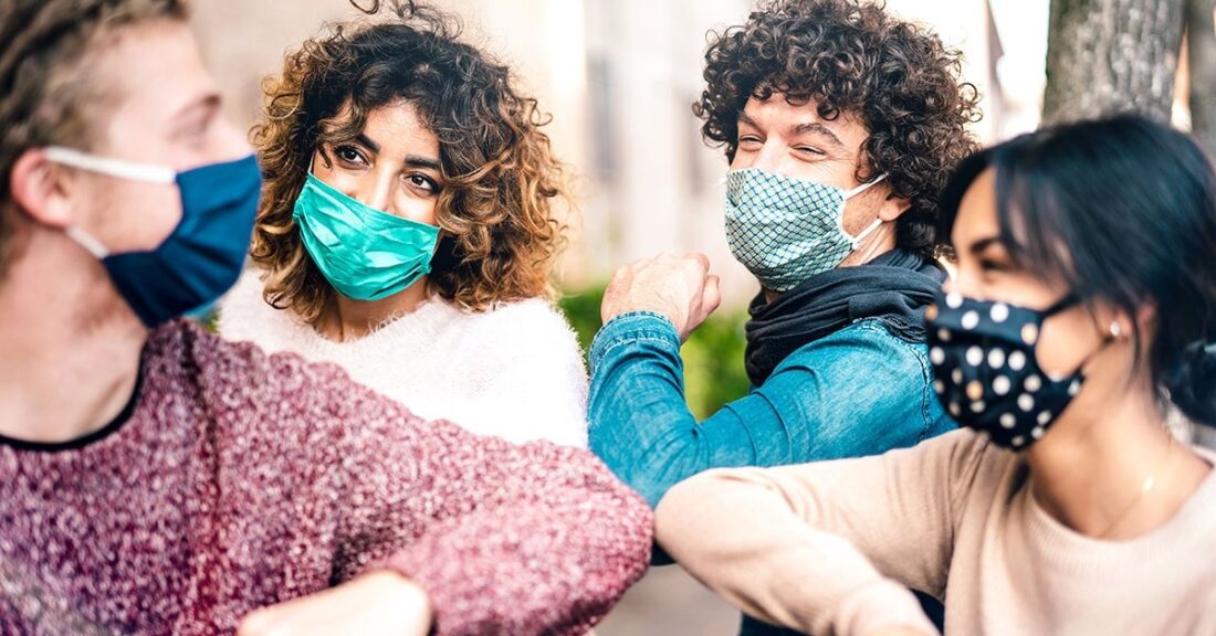 Four young people—two young white males and two young Brown females—gather outdoors, all wearing masks. They form a tight circle, each with one elbow raised and bent and pointing toward the center.