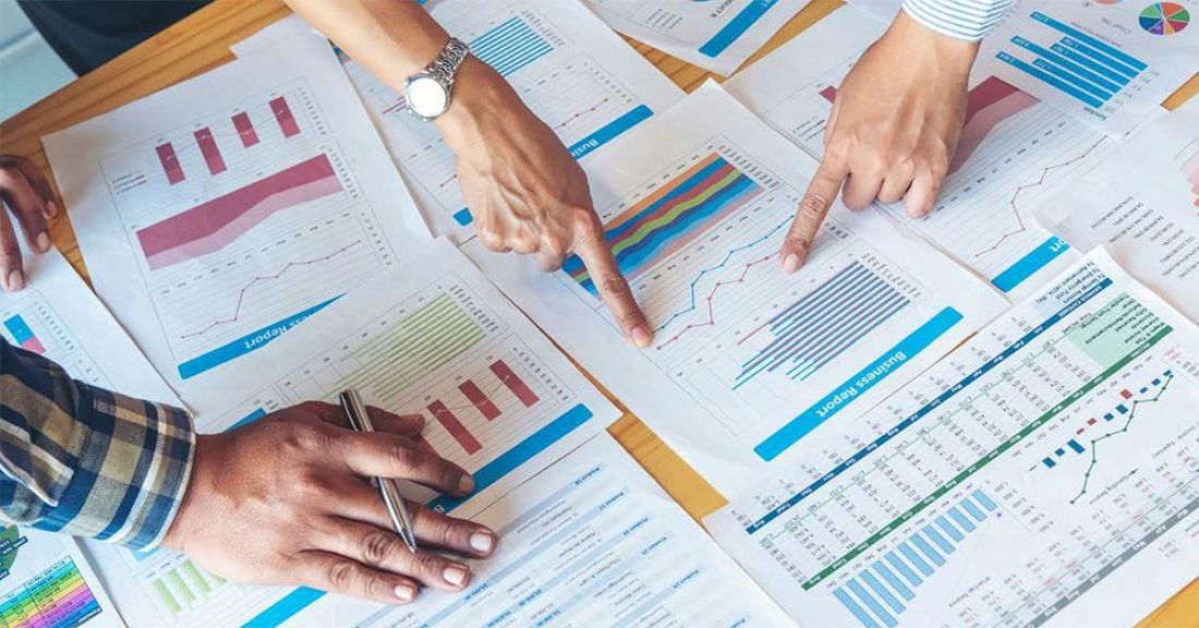 The image depicts an overhead view of a table, which is covered with printouts of colorful graphs and charts. Three people — whose faces we do not see — are pointing toward various points on one of the charts.