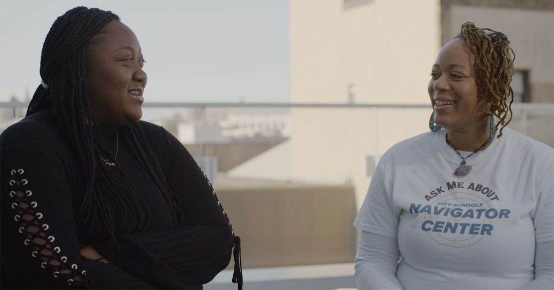 A Black female high school student smiles while conversing with an adult Black woman, who wears a t-shirt that says: “Ask me about City Schools Navigator Center.”