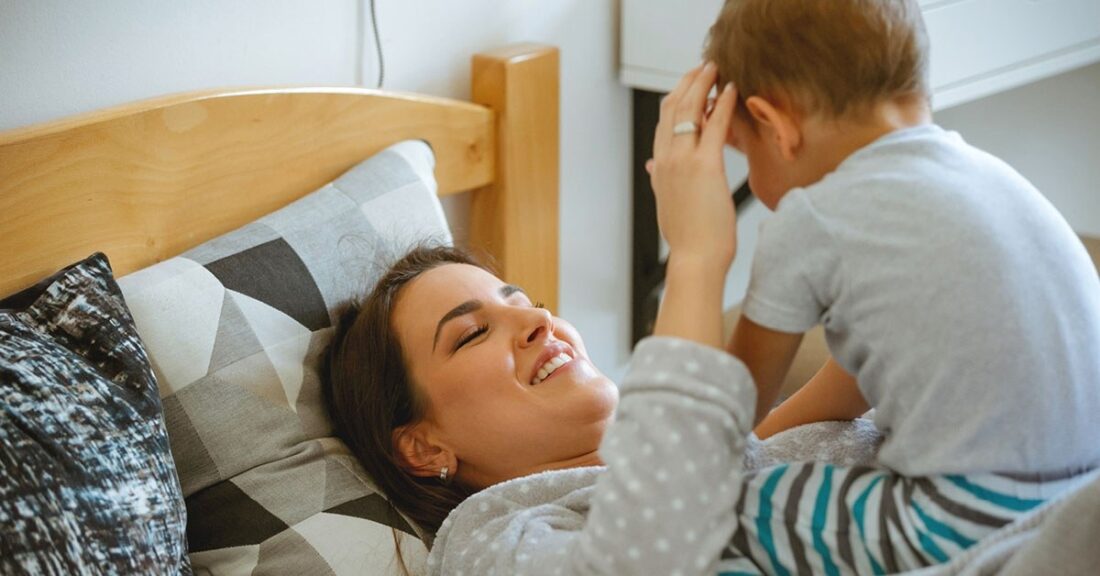 A young brunette woman lies on a bed—smiling, while playing with a toddler who sits on top of her.