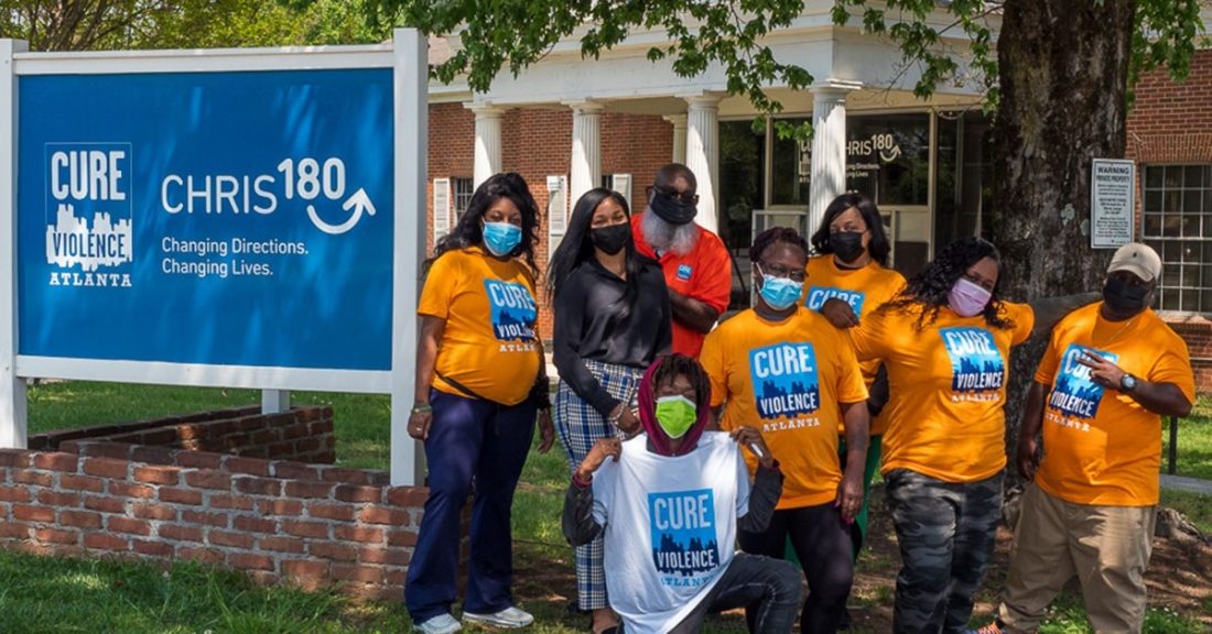 A group of Black men and women gather in front of an office building. They are standing next to a sign for “Cure Violence Atlanta” and CHRIS 180. The adults pictured are all wearing surgical masks, and t-shirts promoting Cure Violence Atlanta.