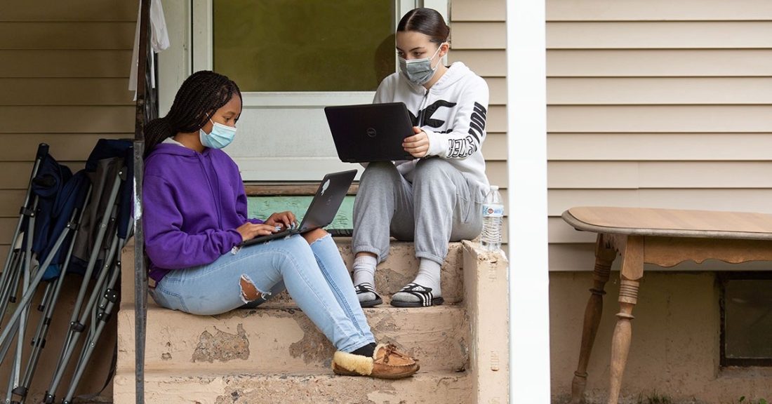 Two teenage girls, one Black and one white, sit on the front steps of a house. They each wear hoodies and surgical masks, while balancing a laptop on their laps.
