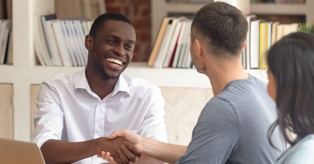 A young Black man wearing a white button-down shirt smiles while shaking hands with a white man, whose back is to the camera. The two men are seated, indoors, and there is a shelf filled with books behind them.