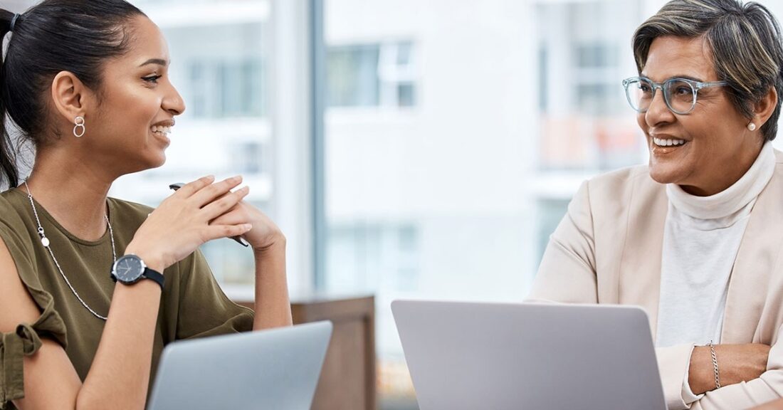 A young Brown woman sits with an older Brown woman, while smiling and conversing. The two are shown in a workplace environment with laptops in front of them.