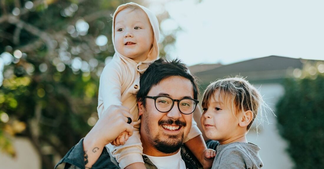 A young father smiles while standing outside with his two children. One toddler-aged child is in front of him, while his younger child perches on his shoulders.