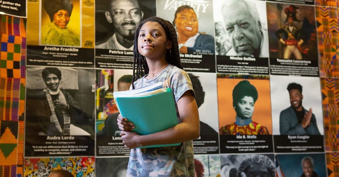 A young Black girl, of approximately middle school-age, stands proudly in front of a mural of Black history-makers, such as Aretha Franklin and Ida B. Wells. She carries a stack of school books in her arms.