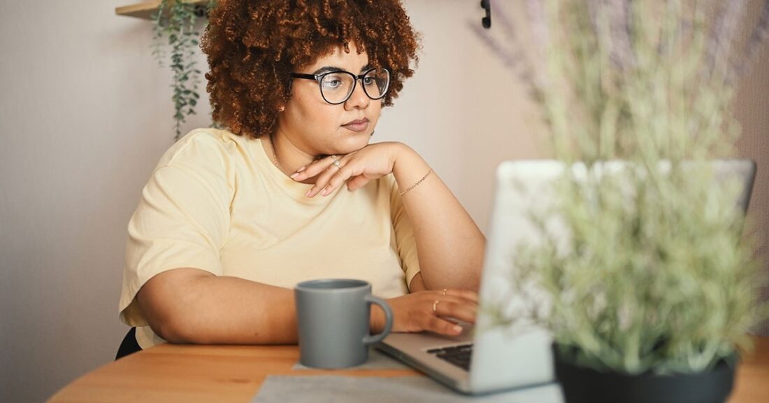 A young Black woman, with natural hair and dark-rimmed glasses, stares intently into her laptop. She rests her chin on her hand.