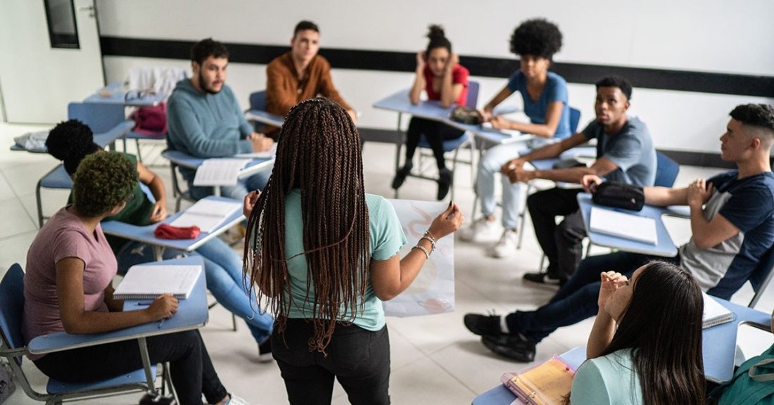 A group of Black and Brown young people gather in a classroom setting. They are seated at desks—in a circle formation. In the middle of the group, a young Black woman holds a visual aid while leading a presentation.