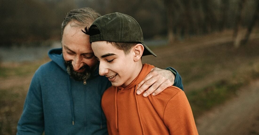 A father and his teenage son—both wearing hoodies—walk together in an outdoor setting. The father’s arm is around his son’s shoulders and the two are smiling.