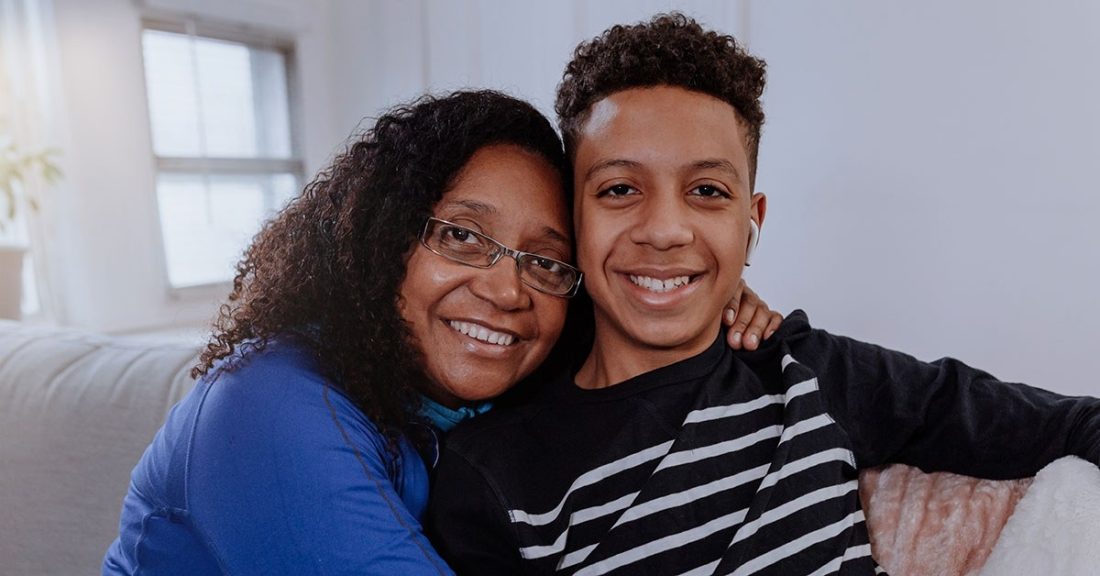 A Black mother and her teenage son embrace, while smiling.