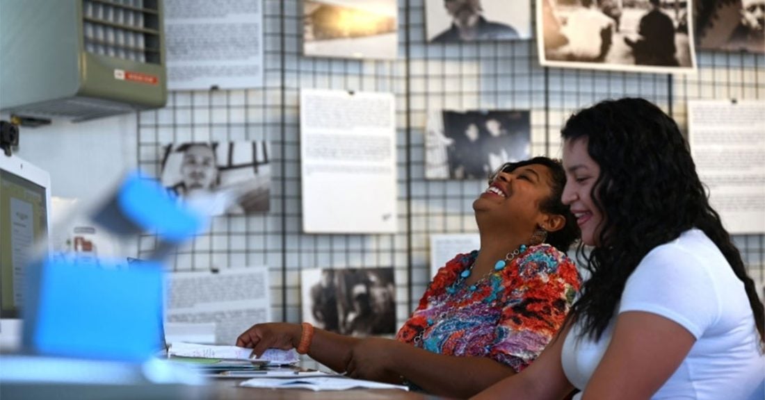 Two young women of color sit side-by-side in an office setting. The two are smiling and laughing; behind them, photos and typed documents are posted on the wall.
