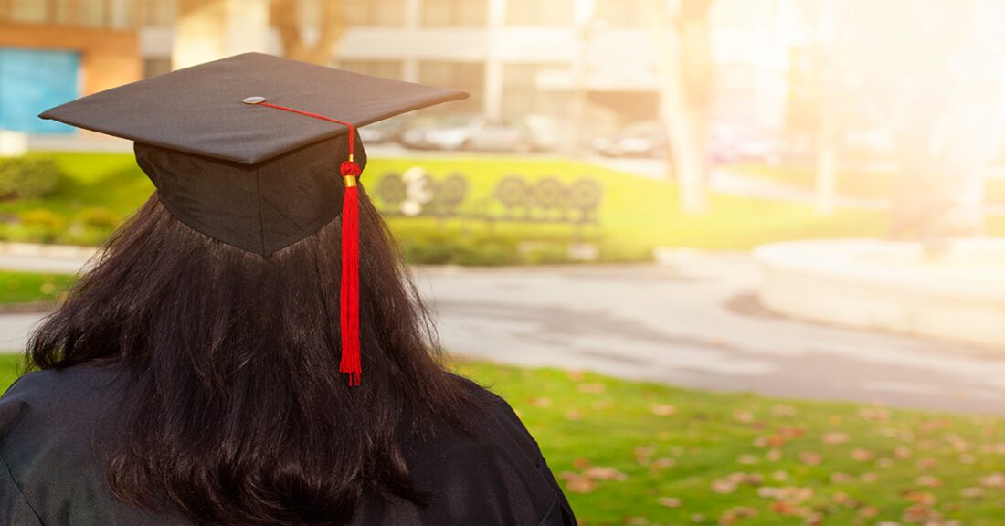 A young black woman wearing a graduation cap stands with her back to the camera. She wears a graduation cap and gown and looks off into a sunny quadrangle.