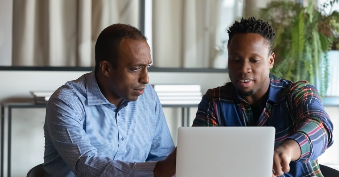 Two Black men sit in front of a laptop. One points at the screen, discussing strategy and sharing ideas.
