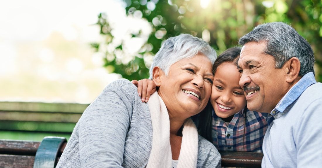 A young Hispanic girl enjoys a day outdoors with her grandparents. They hold each other in a tight embrace. All are smiling.