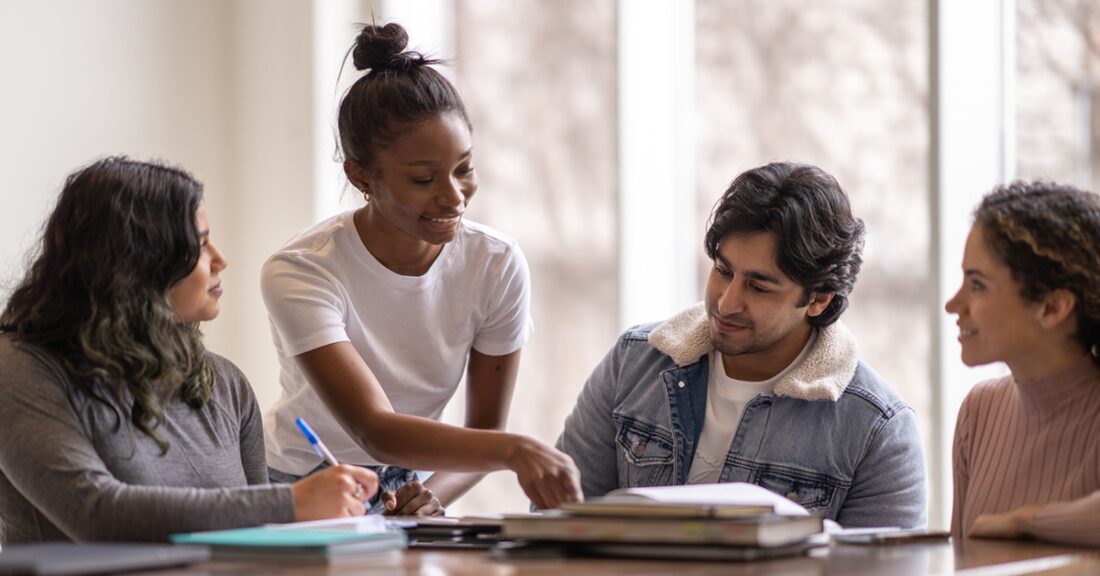 A multiracial group of young people gather around a table, working together. One, a young Black woman, stands, pointing to something in a book. Three other young people sit and smile in agreement with her.