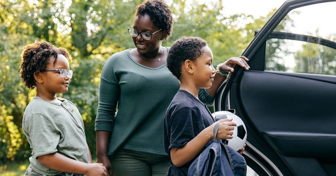 A young Black mother helps load her two small children—one of whom carries a soccer ball—into their car.