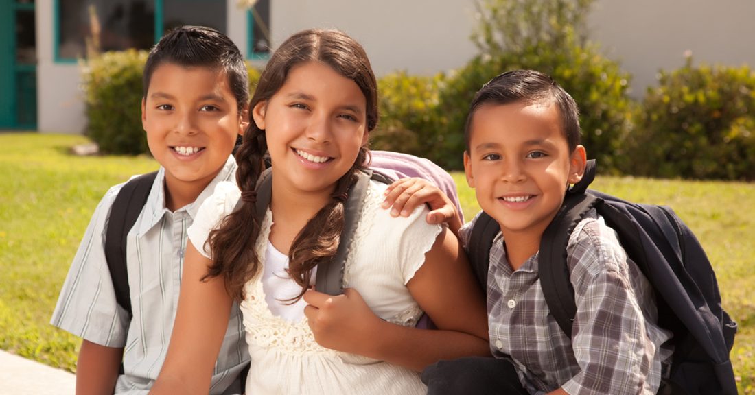 Three Hispanic siblings sit in front of a grassy knoll. They beam at the camera, wearing backpacks.