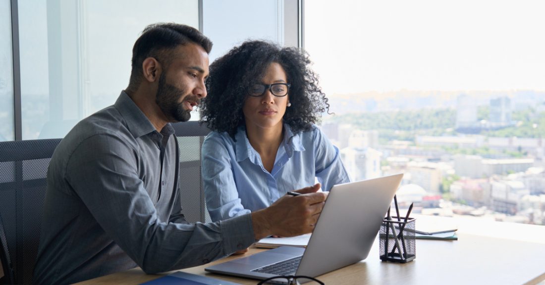 Indian male consults with his female African American colleague using a laptop, sitting in a modern office near a panoramic window.