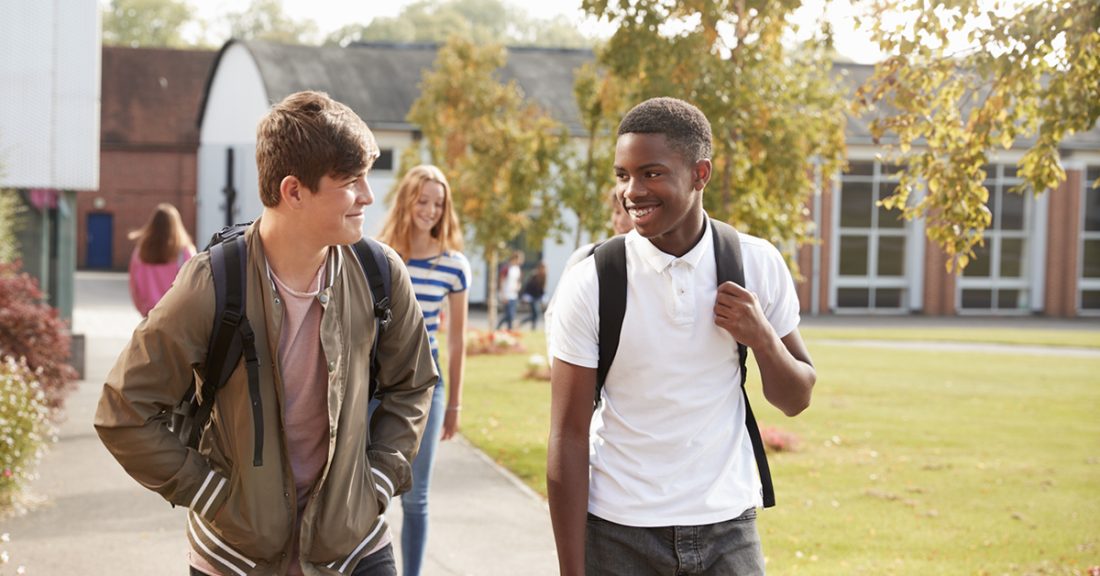 Two middle-school-age boys wearing backpacks — one white and one Black — walk together on a school campus.