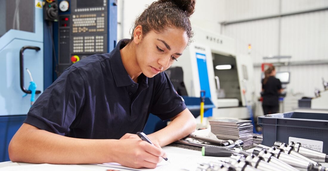 A young woman of color wearing a navy blue polo shirt sits at a workspace — pen in hand — presumably participating in an apprenticeship.