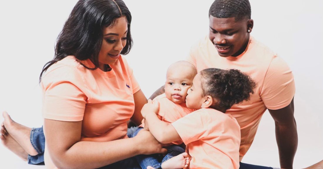 Khalil Peters and his family sits for a photo, wearing matching peach-colored shirts. His wife holds their baby as their oldest daughter kisses the baby. Khalil smiles down at them.