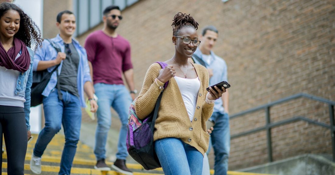 A woman of African descent walks down the steps of campus with more students walking behind her. She is smiling as she carries a backpack around one shoulder and is holding a phone in her hand.