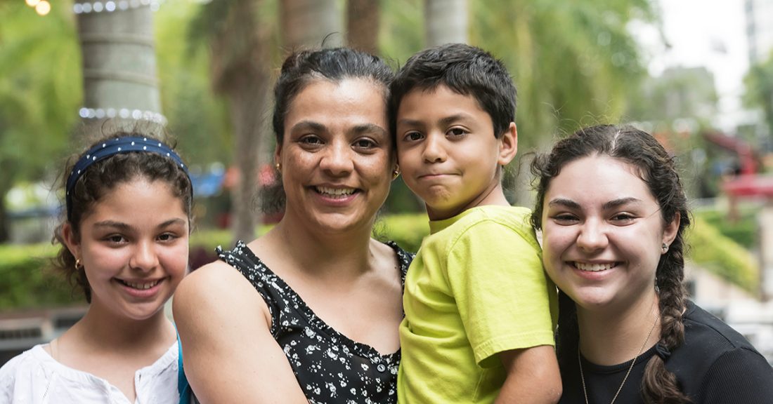 A Latino mother holds her young son; she is flanked by her two middle-school-aged daughters. Everyone is smiling into the camera.