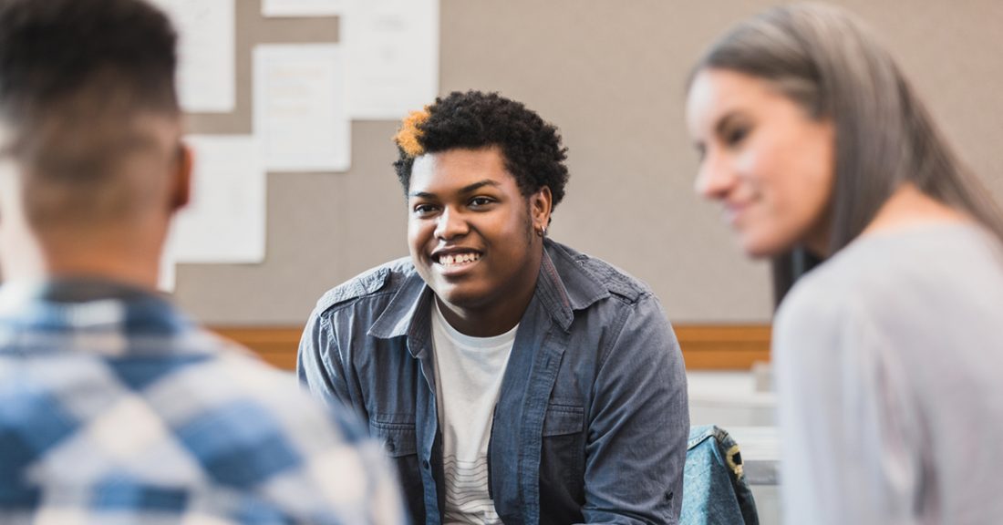 An unrecognizable white female teacher sits in the right portion of the frame. She's smiling at a boy who is obscured in the foreground. In the middle of the image, in focus, a Black teenage boy smiles and welcomes the student in the foreground.