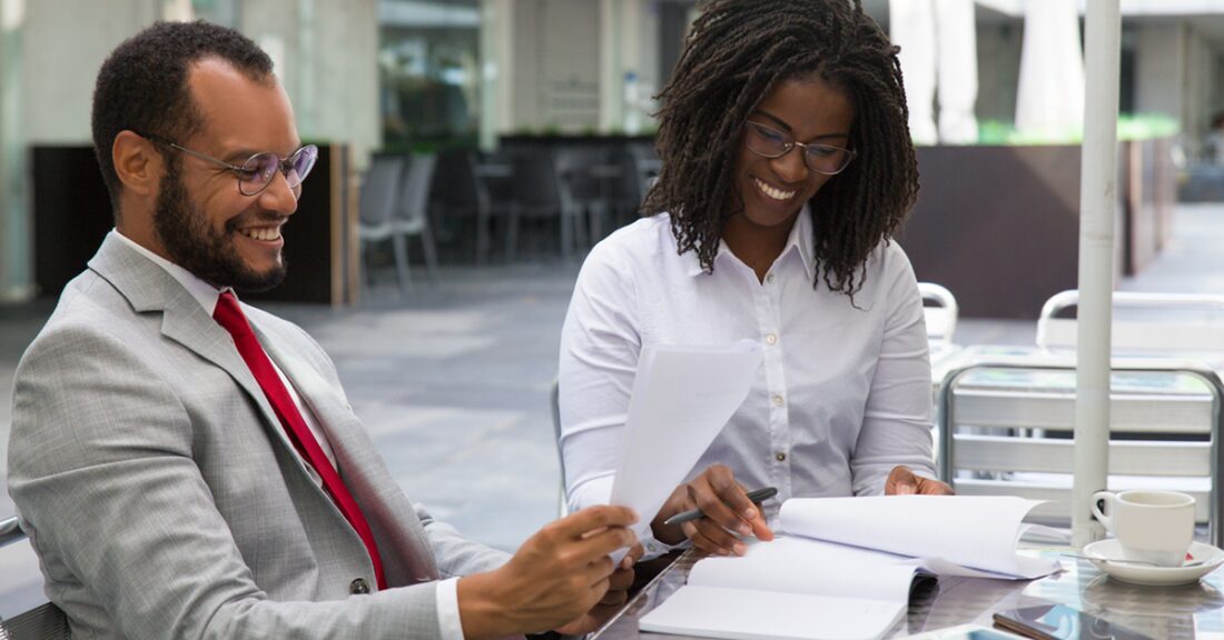 A Black businessman and a Black businesswoman study reports in a coffee shop. As they review their paperwork, they talk, smile and laugh.