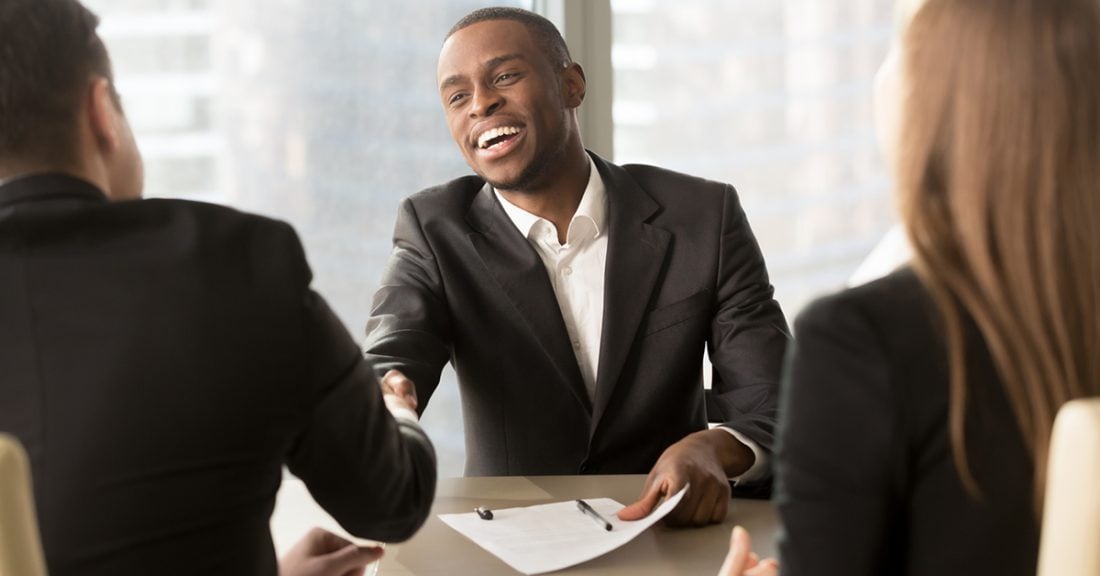 A Black man smiles while sitting across from two white hiring managers, whose backs are to the camera. The Black man shakes the hand of the male hiring manager, while holding his resume in his other hand.