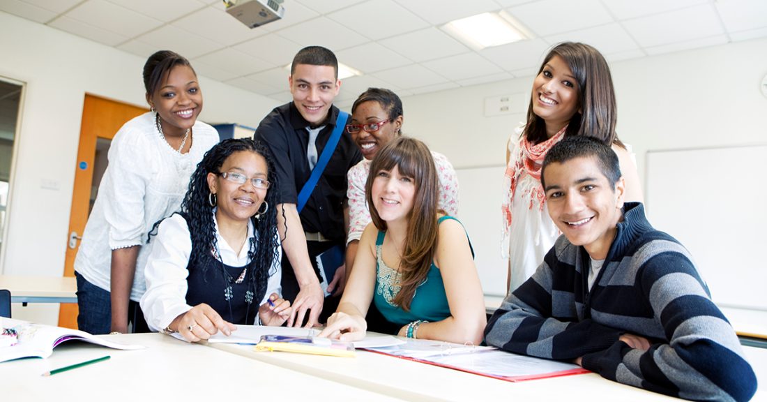 A multicultural group of young people sit and stand around a table, working on a project. They smile confidently at the camera.