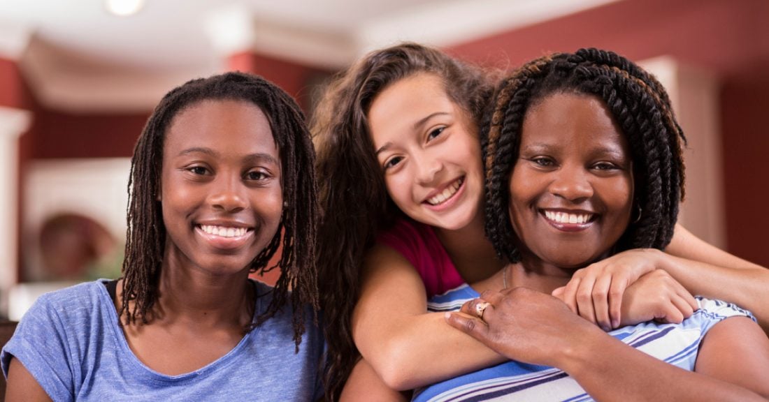 A happy family sits together. They smile at the camera. On the left side of the frame is a Black teen girl. In the middle of the frame is a multiracial teen girl, who hugs a Black woman to her right.