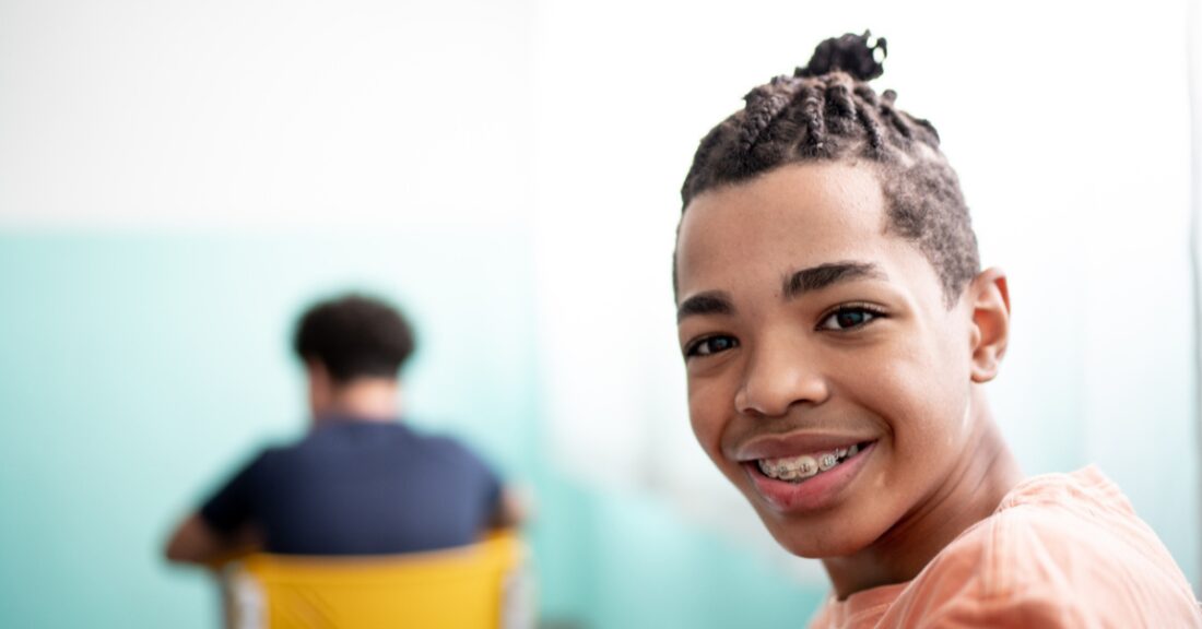 A young, Black teenage boy smiles into the camera, while sitting in a classroom setting. The boy wears braces and his hair is styled in braids.