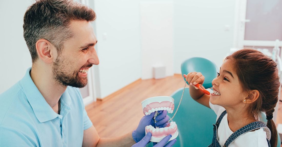 A male dentist smiles while showing a young girl how to properly brush her teeth — using a handheld dental model and a toothbrush.