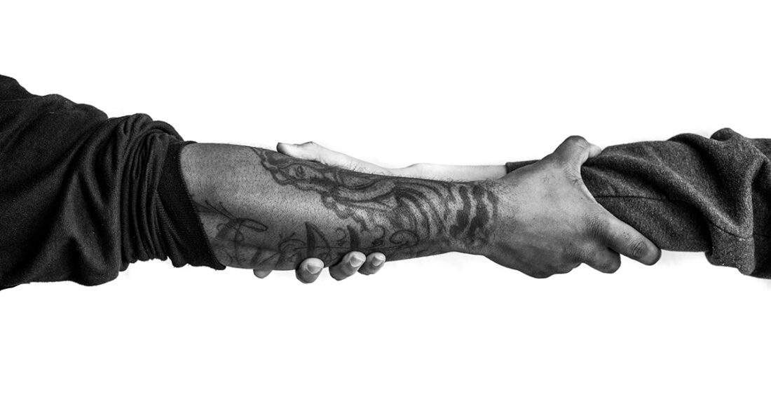 Two people grasp arms just beyond their wrists; one arm shows a tattoo.